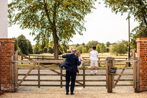 Wedding guest carrying a child while walking toward a wooden farm gate overlooking a scenic countryside landscape.