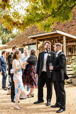 Wedding guests in formal attire socializing outdoors near a rustic barn venue on a sunny day.
