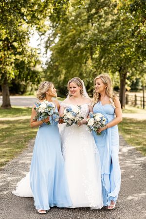 Bride and two bridesmaids in light blue gowns walking along a tree-lined path with bouquets.