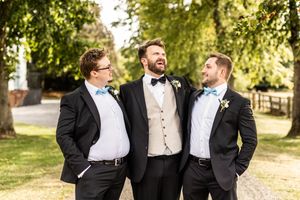 Groom and two groomsmen in black tuxedos with blue bow ties laughing together outdoors.