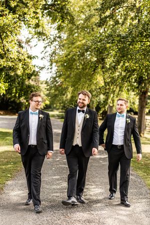 Groom and groomsmen in black tuxedos with blue bow ties walking along a sunny tree-lined path.