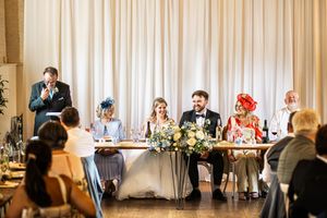 Wedding party seated at head table during reception speeches with guests listening attentively.