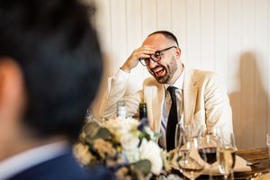 Laughing wedding guest in beige suit and glasses reacting to humorous speech at reception.
