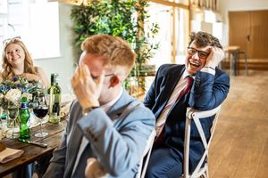 Two male wedding guests laughing and covering faces during amusing wedding speech at reception.