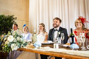 Bride and groom smiling at head table during wedding reception speeches with family seated nearby.
