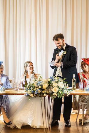 Groom delivering wedding speech at head table while bride smiles beside floral centerpiece.
