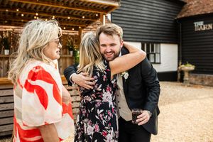 Groom in suit warmly embracing wedding guests outside rustic barn wedding venue.