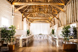 Elegant barn wedding reception with long tables, white draping, fairy lights, and lush greenery in a rustic timber-framed venue.