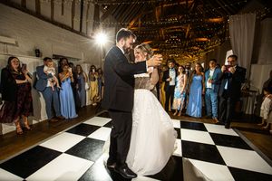 Newlyweds sharing first dance on checkered floor surrounded by guests in a barn venue.