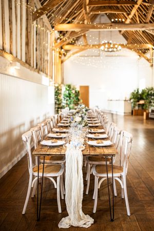 Rustic barn wedding reception featuring a long wooden table with white fabric runner, floral centerpieces, and fairy lights overhead.