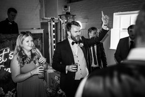 Groom in tuxedo standing by photo booth with guests celebrating at wedding reception.