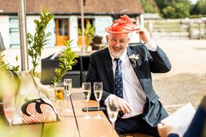 Smiling wedding guest in a navy suit wearing a red fascinator hat while sitting outdoors with champagne glasses.