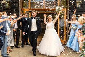 Joyful newlyweds walking through a confetti shower surrounded by cheering guests outside a rustic barn wedding venue.