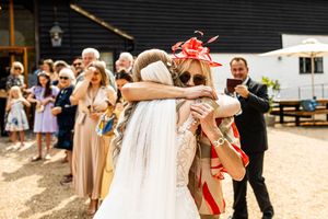 Bride in a lace wedding gown embracing a guest wearing a red fascinator at an outdoor barn wedding celebration.