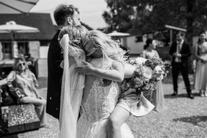Black and white photo of a bride in a lace gown being lifted by the groom during an outdoor wedding celebration.