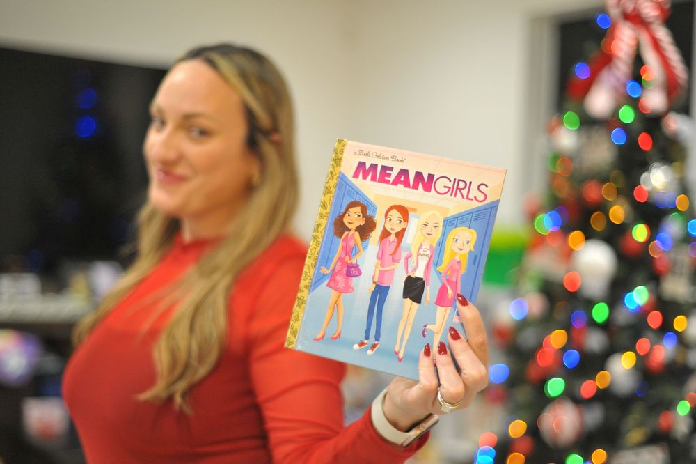 Person holds up Mean Girls DVD and merchandise against festive holiday lights background.