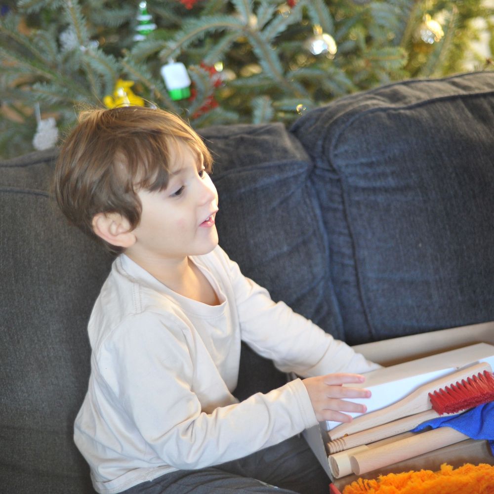 A young student sits on a gray couch with school supplies, with holiday decorations visible in the background.