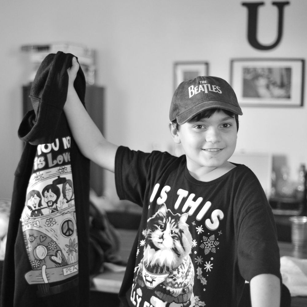 A person holds up a black t-shirt with graphic design while wearing a Beatles baseball cap.