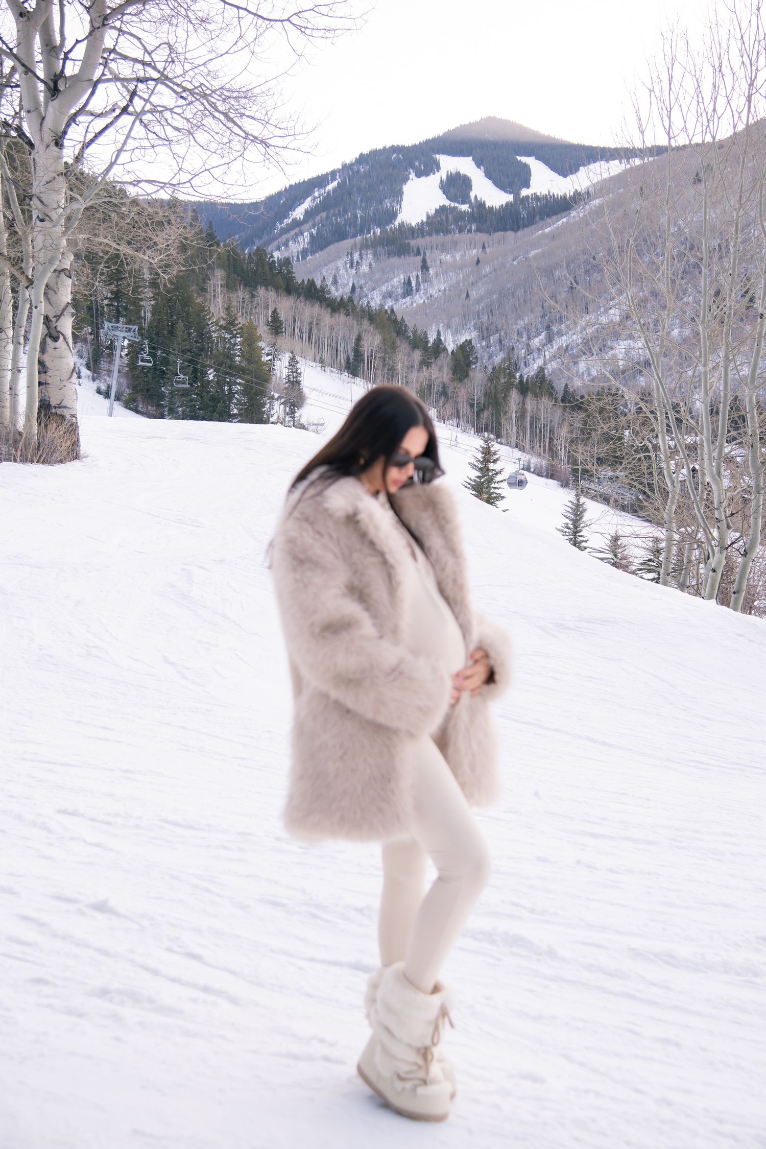 Cute woman in a luxurious beige fur coat poses against a snowy mountain landscape for maternity photo session in Vail Colorado