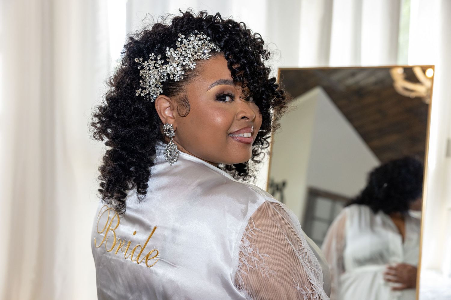 A bride with natural curly hair smiles while getting ready for her wedding day in a white robe.