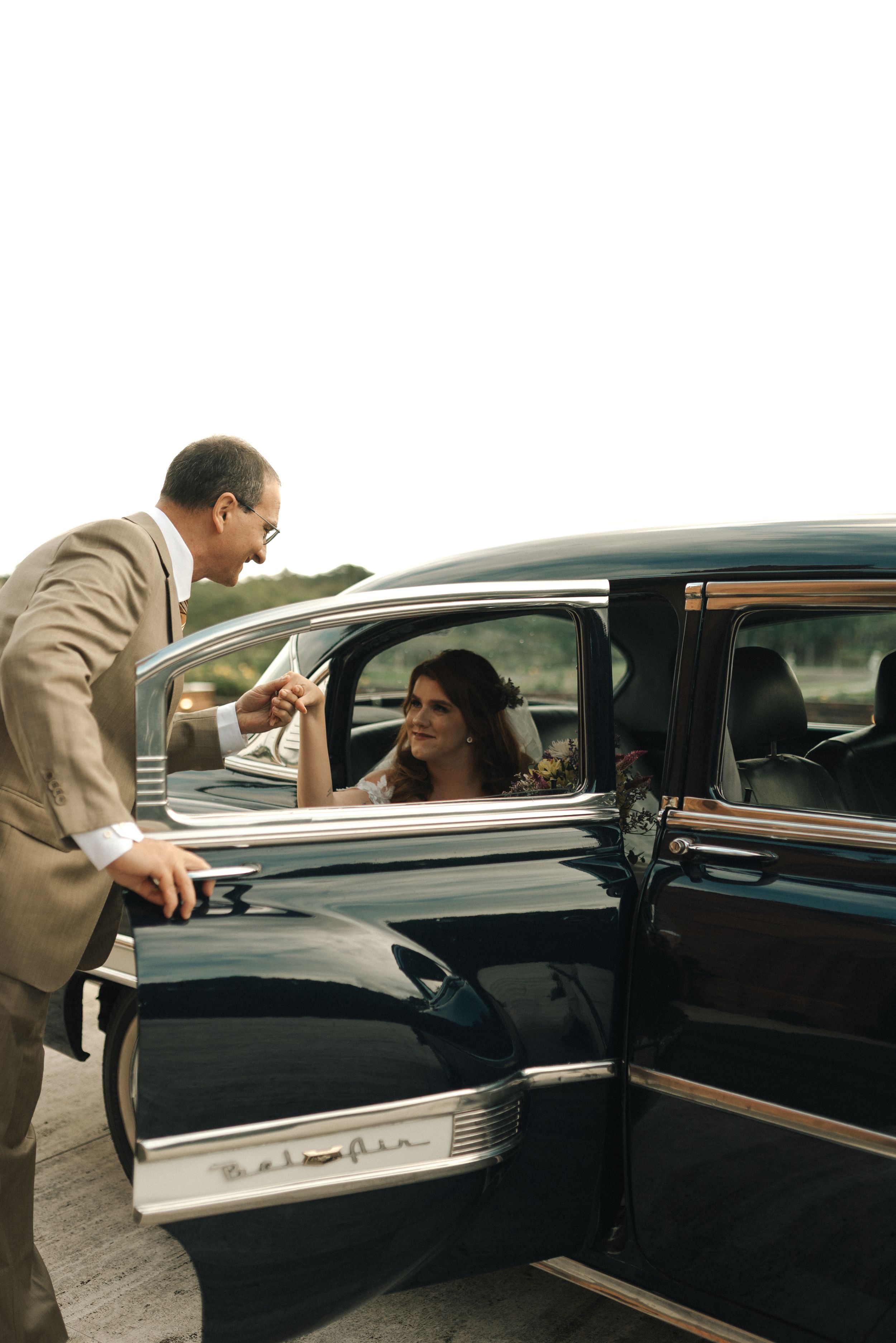 A person in a beige suit leans into a classic car to speak with someone seated inside the vintage vehicle.