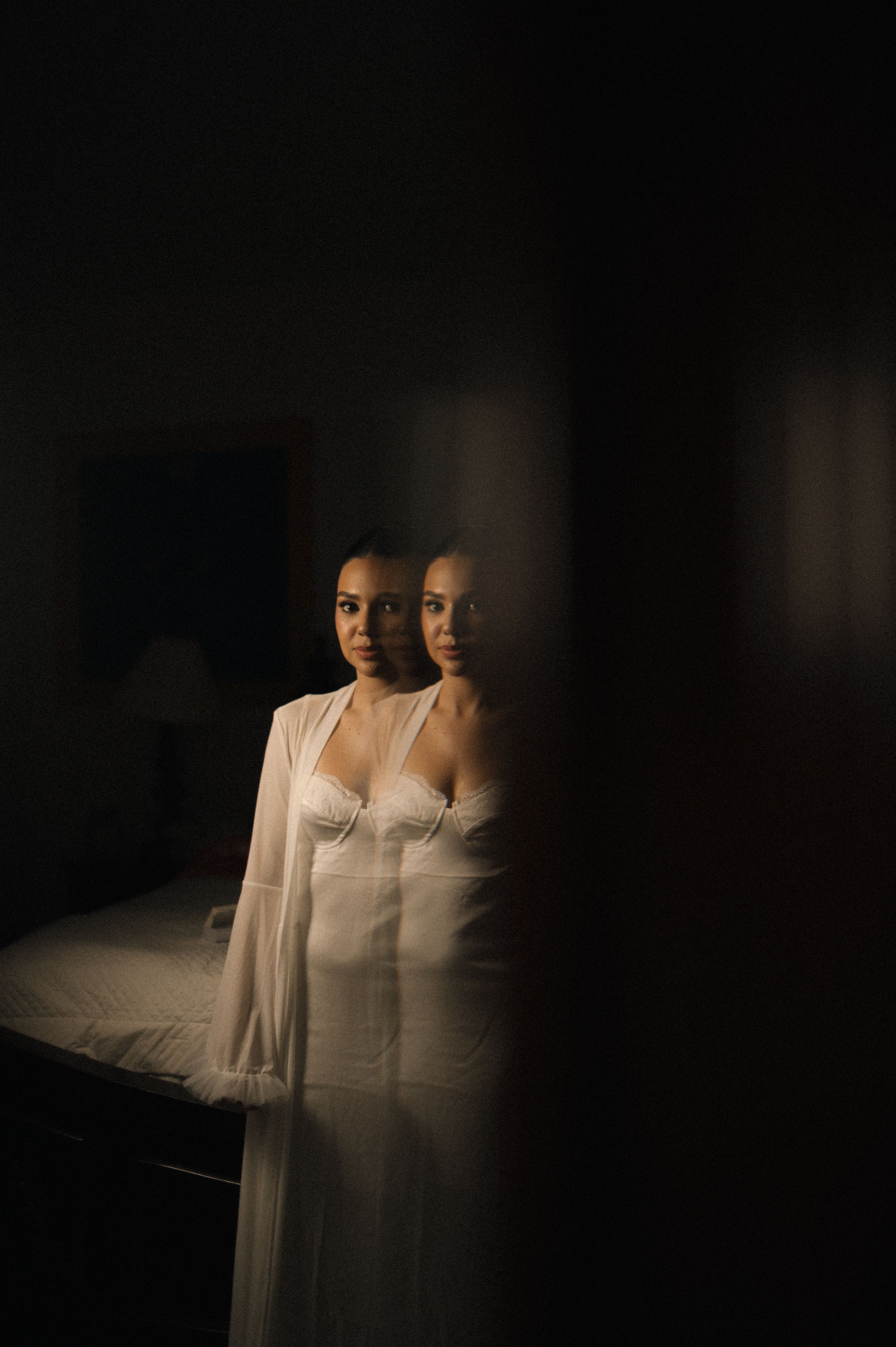 Artistic double exposure portrait in dark moody lighting shows reflection against glass wearing white flowing garments.