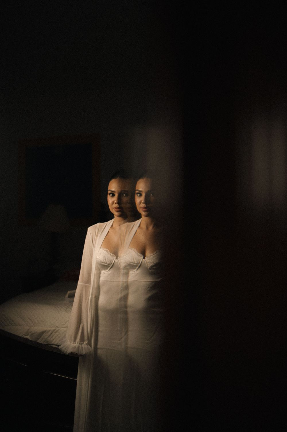 Artistic double exposure portrait in dark moody lighting shows reflection against glass wearing white flowing garments.