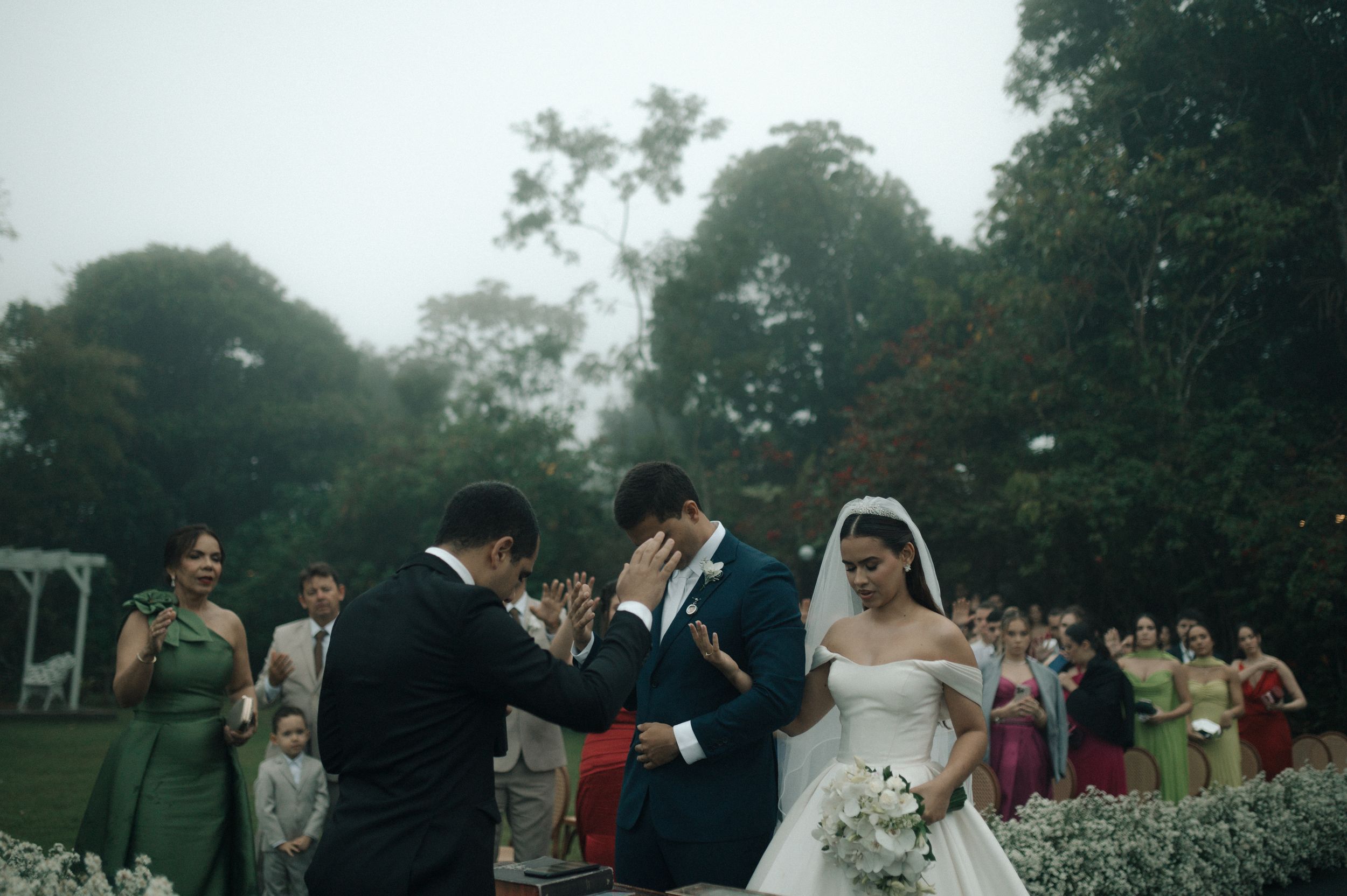 A wedding ceremony takes place outdoors in a garden setting with guests gathered around on a misty afternoon.