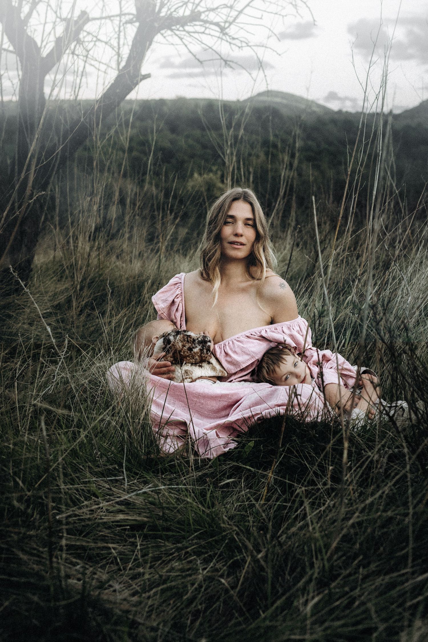 A mother in a pink dress reclines among tall grasses in a moody outdoor photoshoot at dusk. Photographer Javea