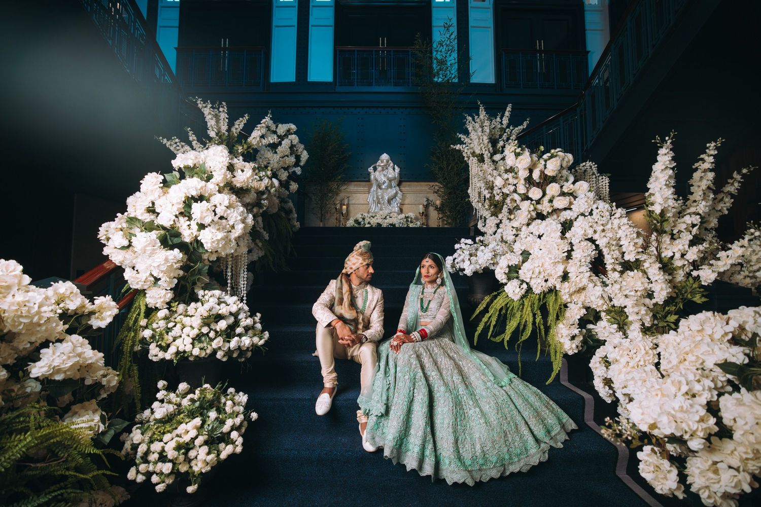 Elegant wedding party poses at night with dramatic lighting and white floral arrangements in a luxurious venue.