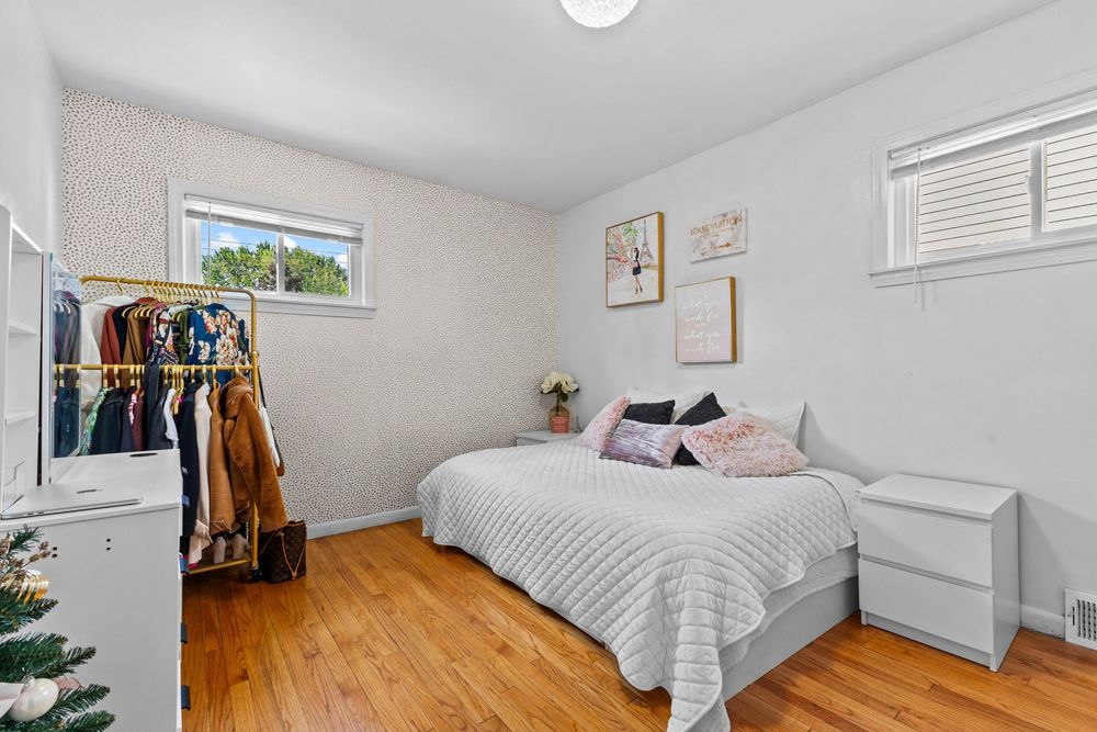 Bright bedroom with hardwood floors, a white bed, open clothing rack, wallpaper accent wall, and natural light from windows.