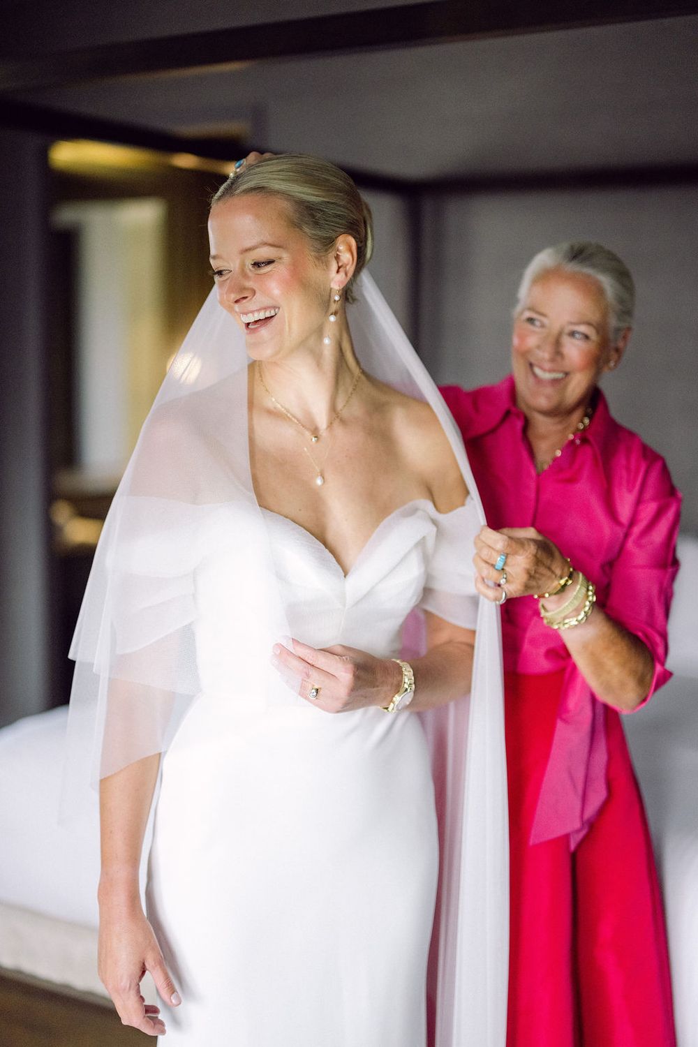 A woman in a white wedding dress stands with someone in a bright pink outfit during wedding preparations.