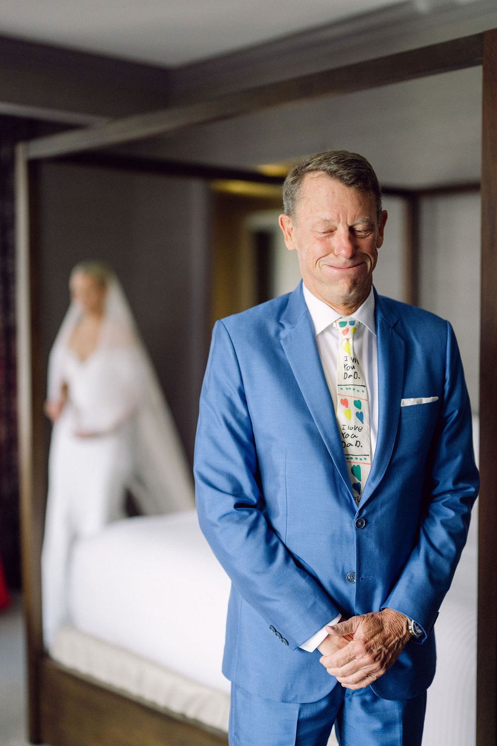 A person in a bright blue suit and patterned tie stands in a room with a bride in white visible in the background.