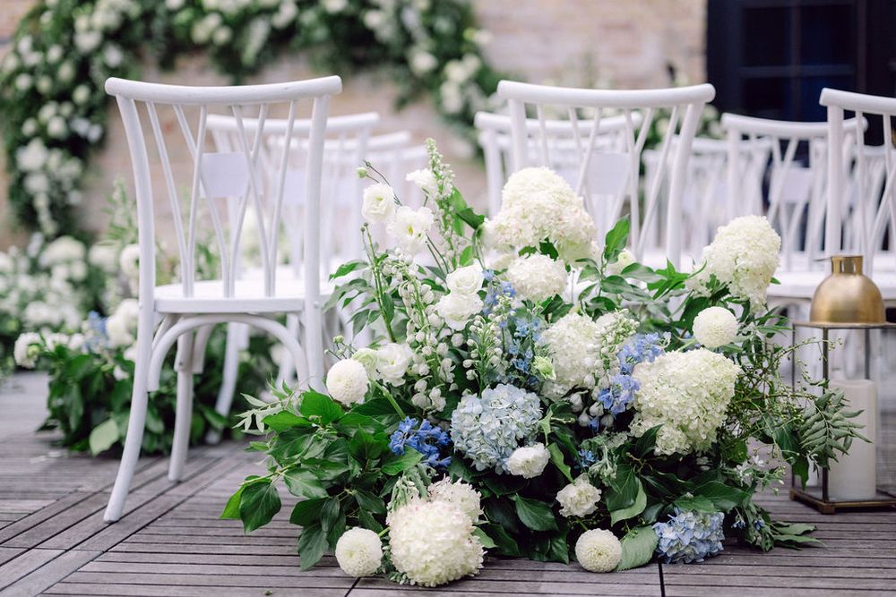Elegant floral arrangements with white hydrangeas and blue flowers line a rustic wooden deck at an outdoor event.