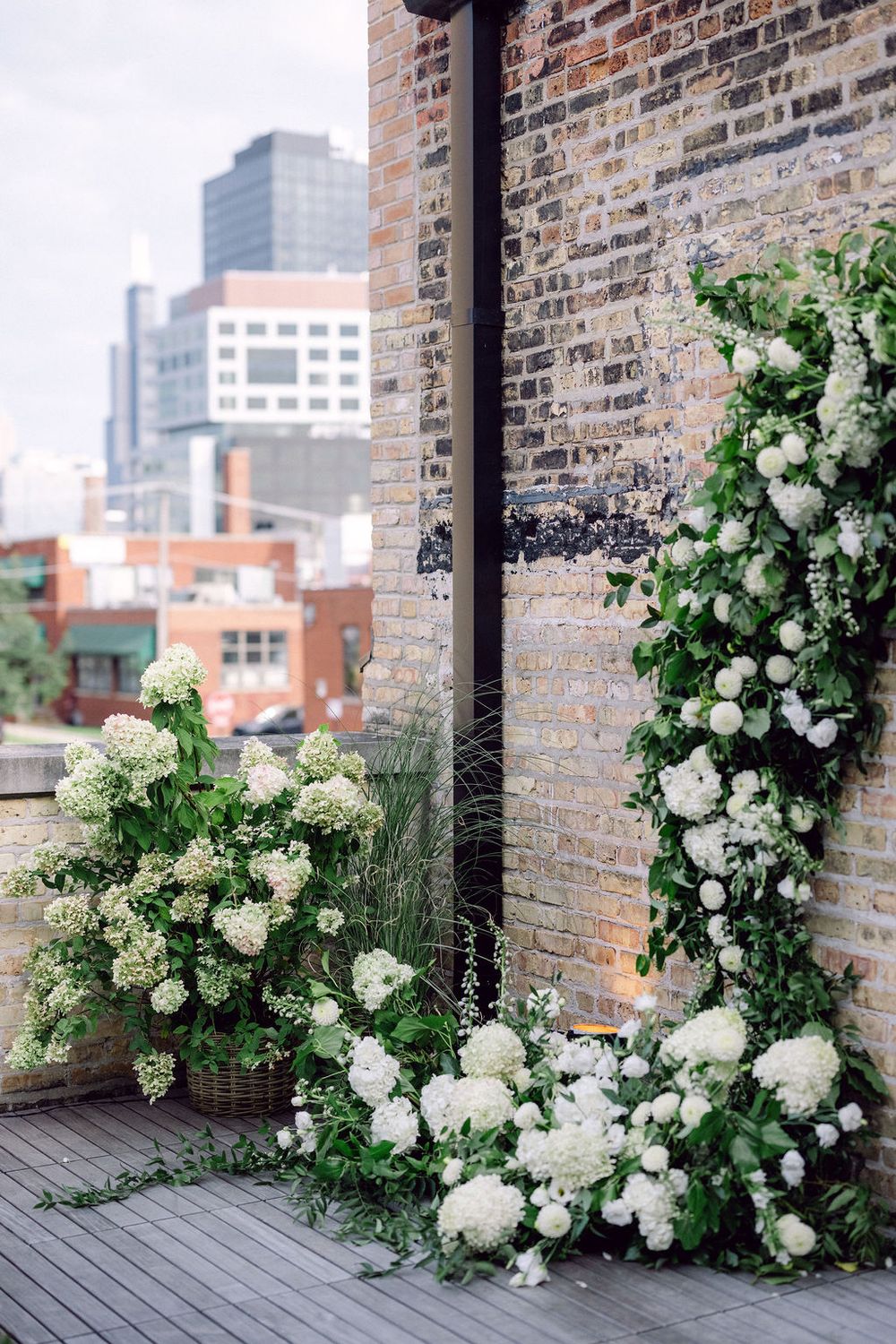 White roses and greenery adorn a brick wall on an urban rooftop terrace with city buildings visible in the background.