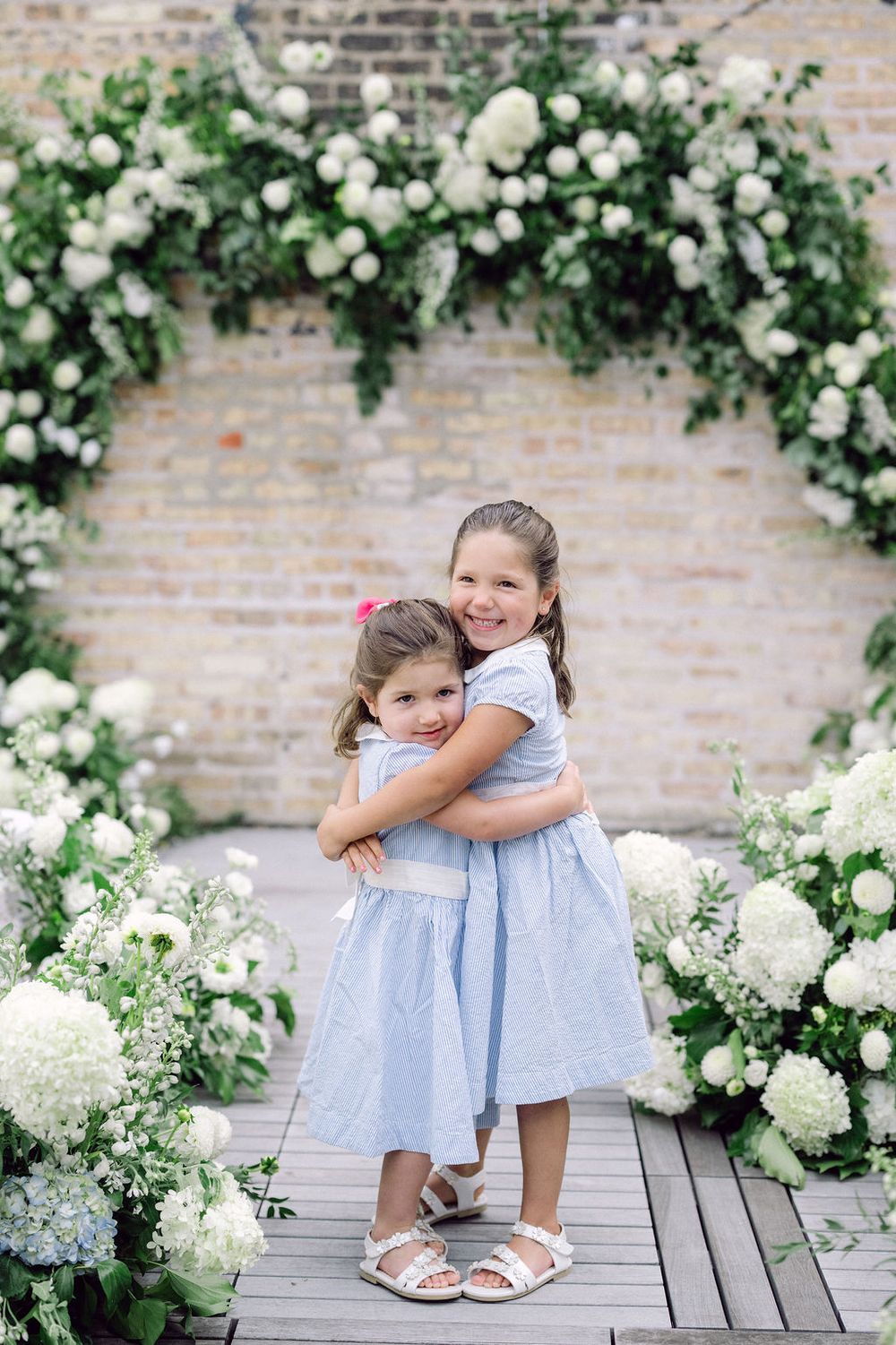 Two young girls in light blue dresses share a sweet moment among white floral arrangements and brick walls.