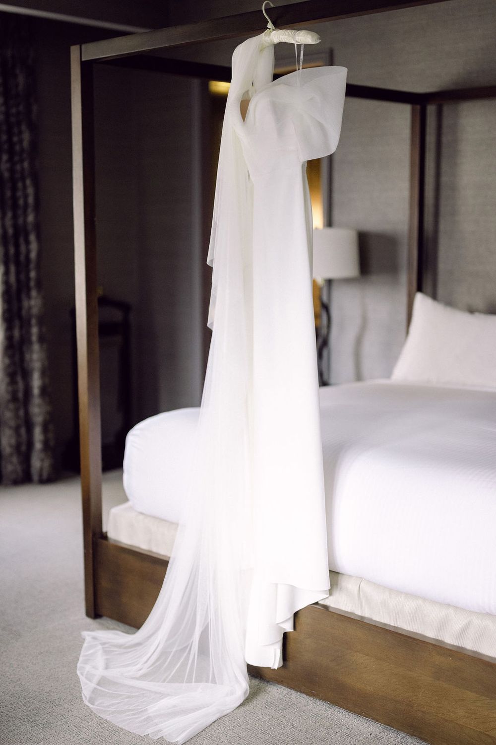 A flowing white wedding dress hangs elegantly in a modern hotel room with dark wood floors and white bedding.