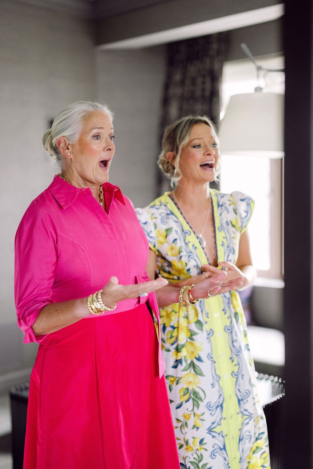 Two women share an excited moment together, one wearing bright pink and the other in a yellow floral print dress.