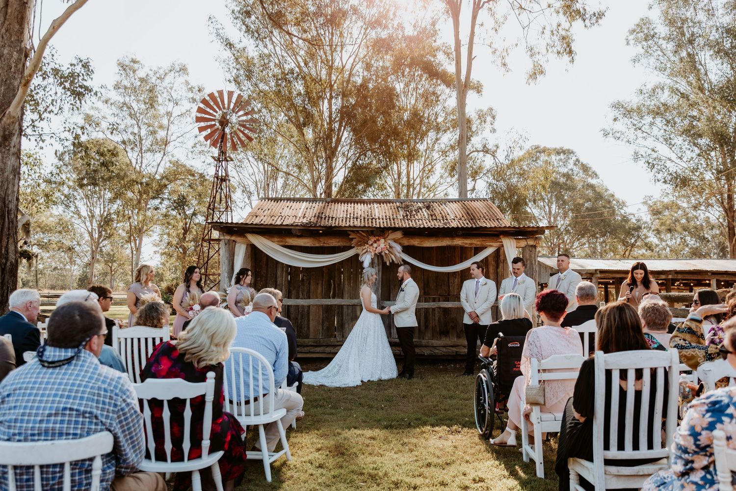 Wedding guests seated at outdoor ceremony with rustic wooden backdrop.
