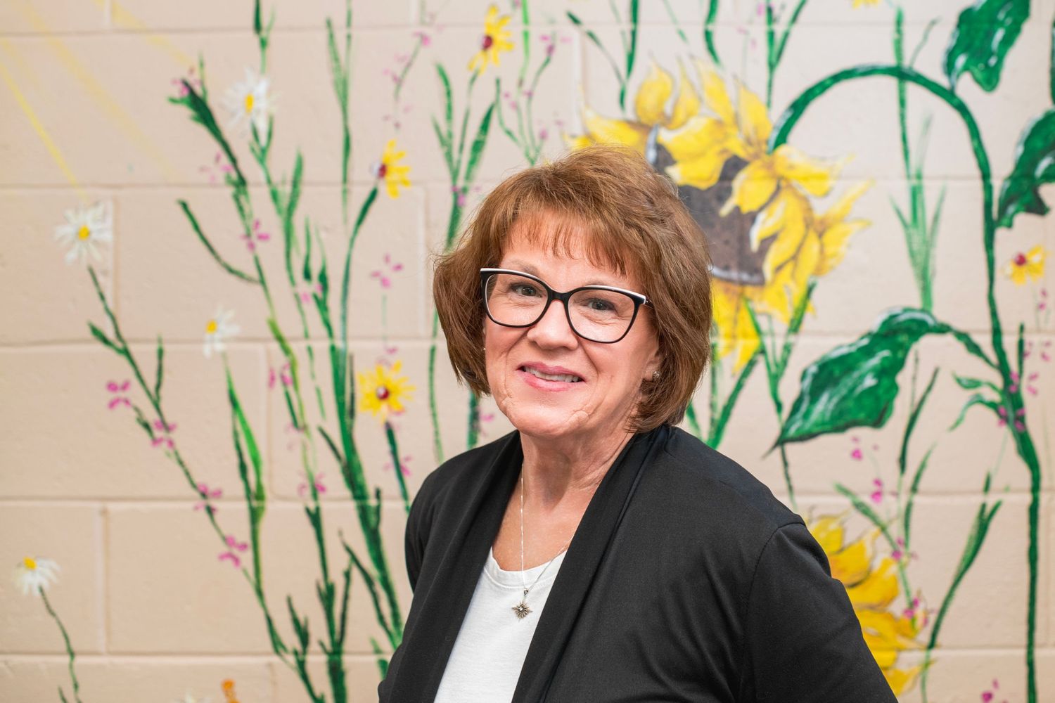 Smiling woman with glasses posing in front of sunflower wall mural.