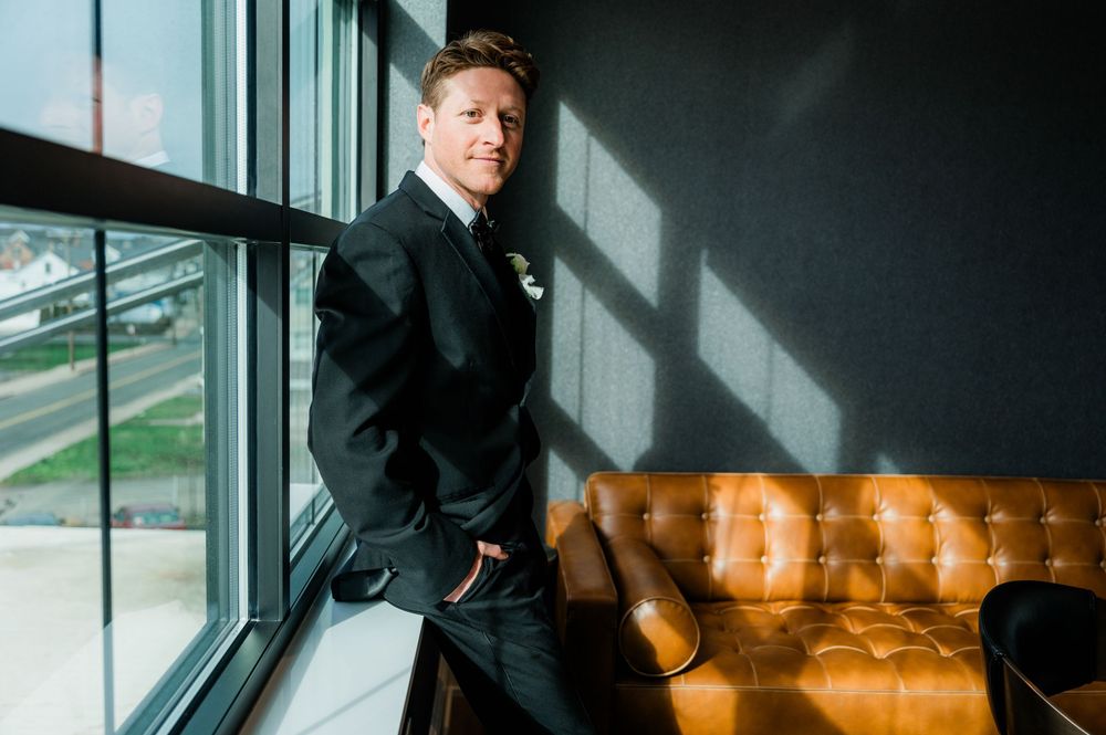 Professional in a dark suit stands by a window in an office building with city views in the background.