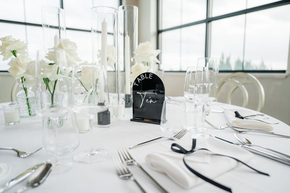 Modern white dining table setup with clear glassware and minimalist place settings near large windows.
