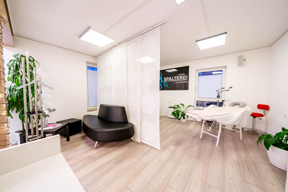 Modern office waiting area with black curved sofa, light wood floors, and potted plants in a bright white space.