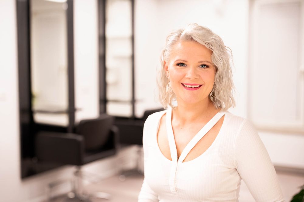 A smiling woman with blonde hair poses in a bright, modern interior space with framed artwork on the walls.