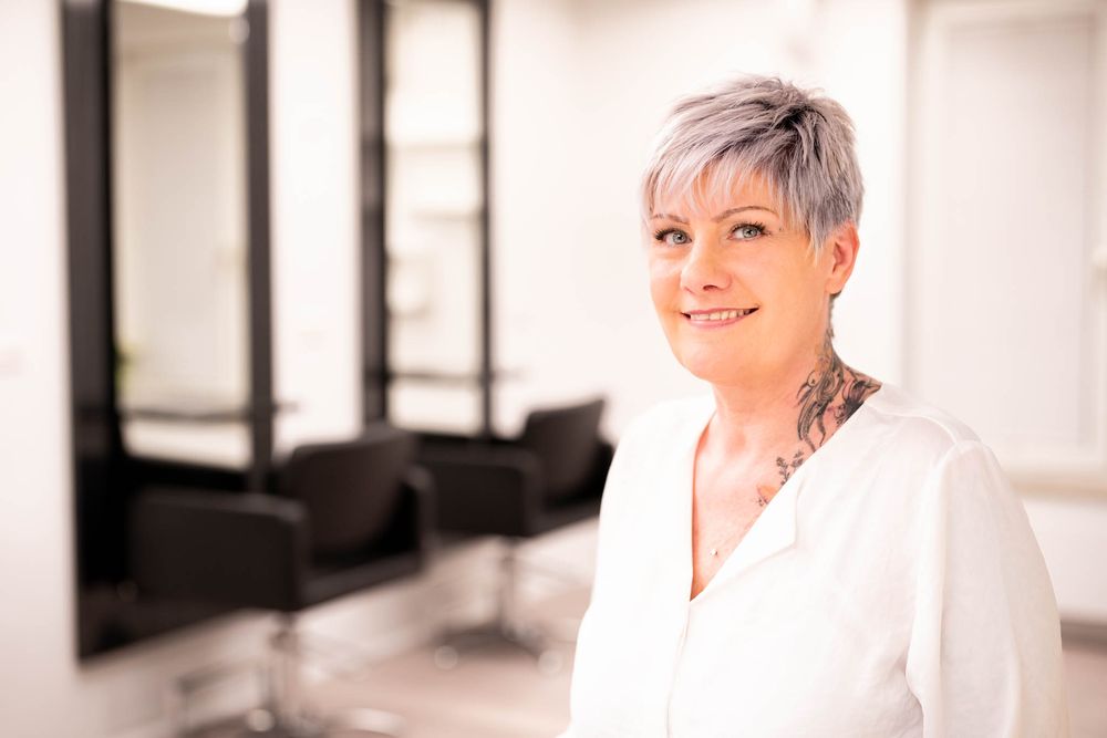 Professional wearing a white button-down shirt smiles warmly in a bright modern office setting.