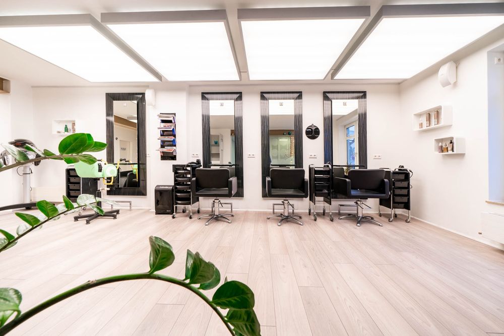 Modern hair salon interior with black styling chairs, large mirrors, and light wood flooring in a bright minimalist space.