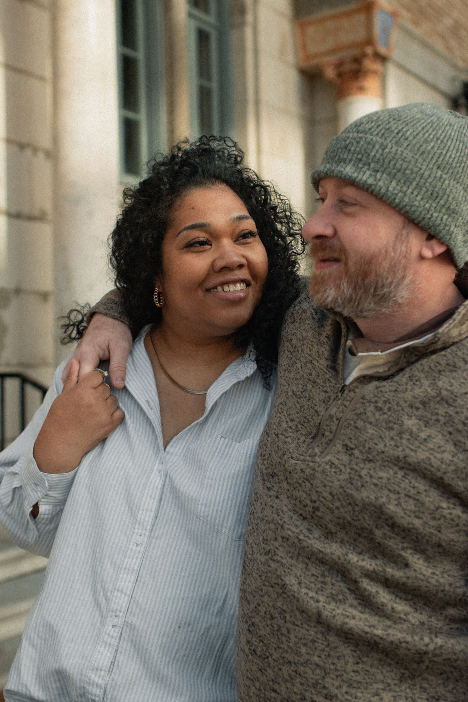 A happy couple embraces and smiles while standing outside a building with architectural details.