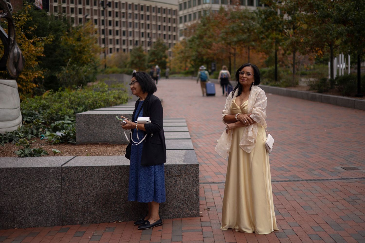Two people in long dresses stand on a brick plaza near garden planters and buildings on an autumn day.