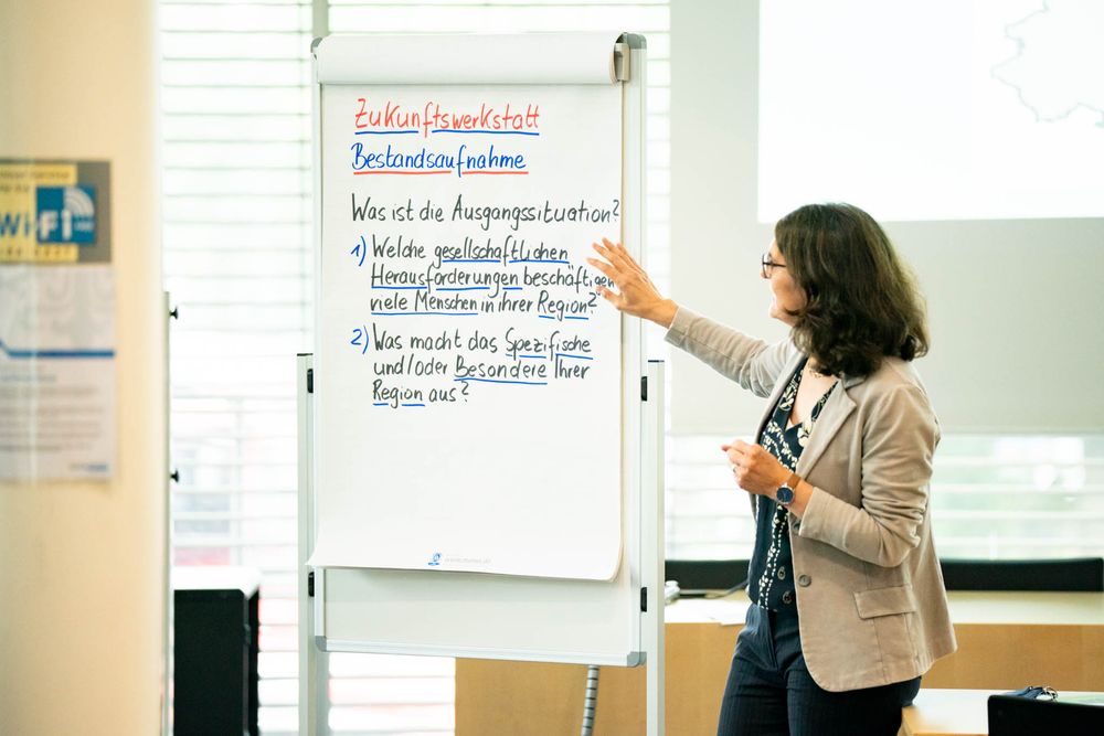 A teacher writes on a whiteboard during a classroom presentation while gesturing with their hand.