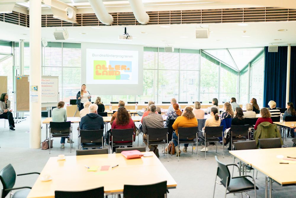 A bright modern classroom with students seated at tables watching a presentation on a projector screen.